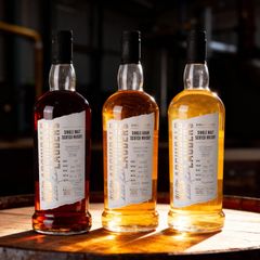 Tre flaskor whisky placerade på ett träfat. Flaskorna har olika nyanser av mörkare brunt och gult.