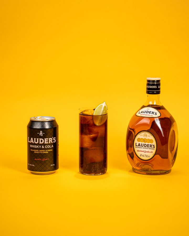 En burk och en flaska Lauder's whisky bredvid ett glas med whisky och cola, dekorerat med en limeklyfta, mot en gul bakgrund.