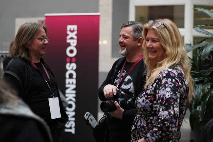 Tre personer, en med kamera i handen, samtalar och ser glada ut.