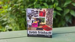 En bok med titeln "Sven Fristedt – mästare i mönster" står på ett bord med gröna växter i bakgrunden.