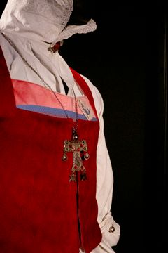 En rött sidenliv med en bröstlapp, matchad med en gammal vit skjorta från sydöstra Skåne, tillsammans med ett dekorativt halsband med ett metallkors.