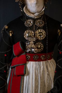 Bild av traditionell folklig högtidsdräkt med dräktsilver. Dräkten har silverdetaljer och ett rött midjeband.
