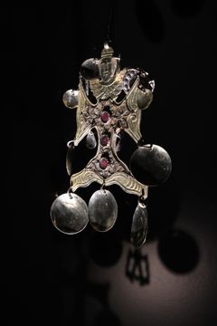 Ett dekorativt kors med ett änglahuvud och vingar, dekorerat med röda stenar och hängande medaljonger.