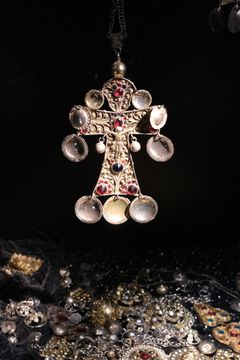 Alt text: Ett dekorativt striglakors i silver dekorerat med röda och vita stenar, hänger över en samling av dräktsilver.