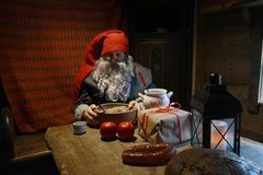 Tomte med skägg, grå yllekläder och röd luva sitter vid ett bord med gåva, korv, ljuslykta och äpplen.