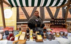 En person står vid ett marknadsstånd med julmat och delikatesser, såsom ostar och konserver, funder en randig markis.