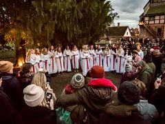 Ett Luciatåg med deltagare i vita klänningar och röda band framför en grupp åskådare utomhus.