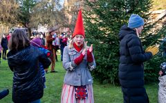 En person i tomtemössa klappar i händerna bredvid en gran, personer rör sig runtomkring. Utomhus.