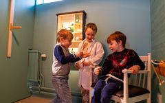 Tre barn leker doktor i en utställning. Ett barn har en vit rock.