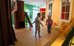 Två barn leker inomhus i en lekutställning med träbyggnader och en rutschkana.