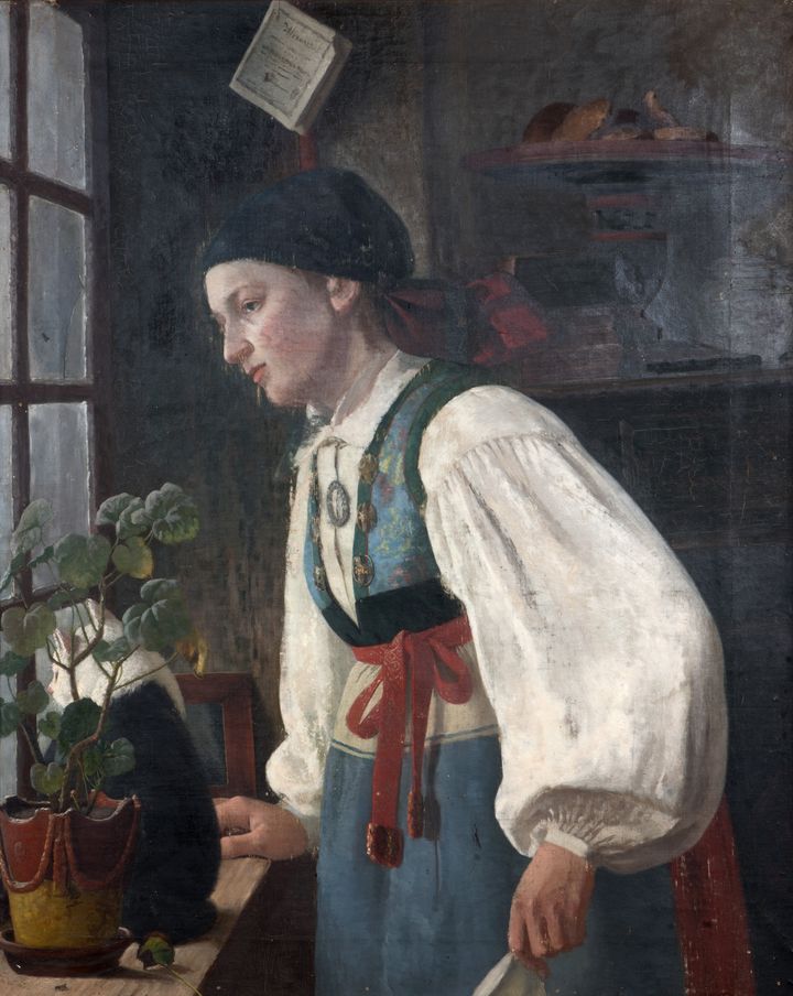 En ung kvinna i skånsk folklig dräkt står vid ett fönster och tittar ut.