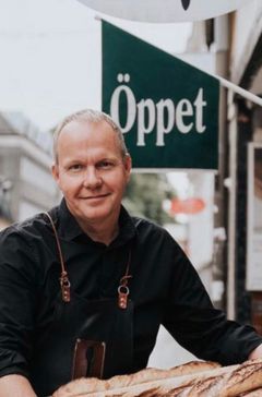 Bild på en man i svart tröja framför en skylt med texten "Öppet", med bröd framför sig.