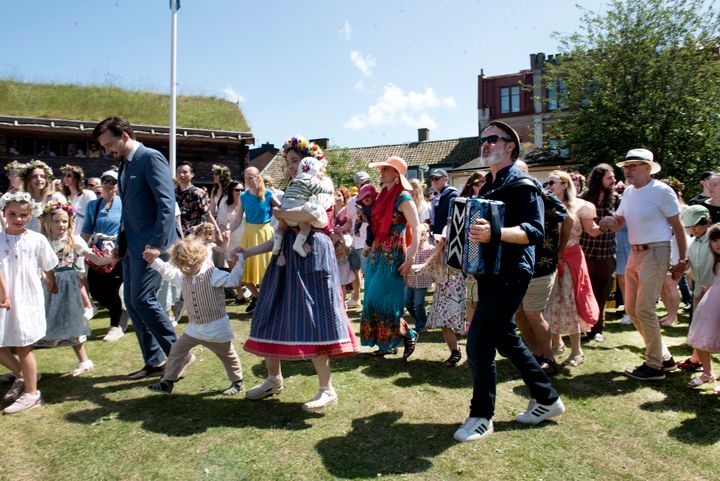 En man spelar dragspel till dansen runt midsommarstången med sommarklädda midsommarfirare.
