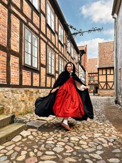 En kvinna klädd i historiska kläder – en röd klänning och en svart cape – är fångad mitt i en rörelse. Hon står på kullersten och är omgiven av korvirkeshus i tegel.