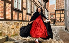 En kvinna klädd i historiska kläder – en röd klänning och en svart cape – är fångad mitt i en rörelse. Hon står på kullersten och är omgiven av korvirkeshus i tegel.
