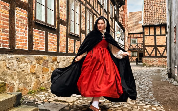 En kvinna klädd i historiska kläder – en röd klänning och en svart cape – är fångad mitt i en rörelse. Hon står på kullersten och är omgiven av korvirkeshus i tegel.