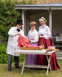 En grupp personer i kläder från 1880-talet står vid ett bord utomhus.