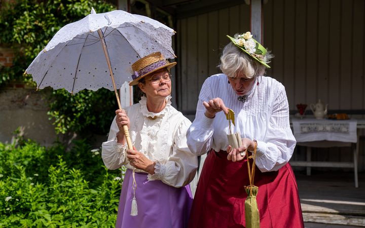 Två kvinnor klädda i 1880-talskläder, en med ett parasoll och den andra tittar på ett föremål i sin hand.