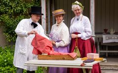 Grupp på tre personer i tidstypiska kläder från 1880-talet står vid ett bord utomhus.