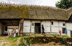 Korsvirkesgård från 1800-talet med halmtak. På gårdsplanen står barn samt en person i tidstypiska kläder.