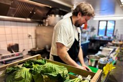 Kock står i köket och förbereder mat. I förgrunden finns en låda med gröna bladgrönsaker.