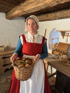 Kvinna klädd i medeltidskläder håller en korg med rotfrukter i en historiskt gestaltad miljö.