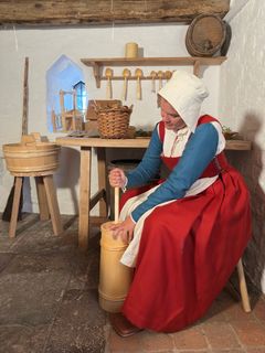 Kvinna i medeltida klädsel sitter och kärnar smör i historisk miljö.