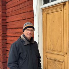 En person i vinterkläder står framför en träbyggnad med en gul dörr.