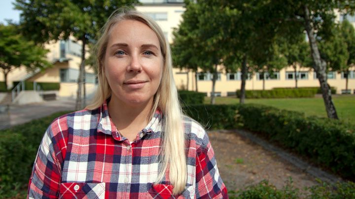 Kvinna med långt blont hår i rutig skjorta står utomhus i en park.