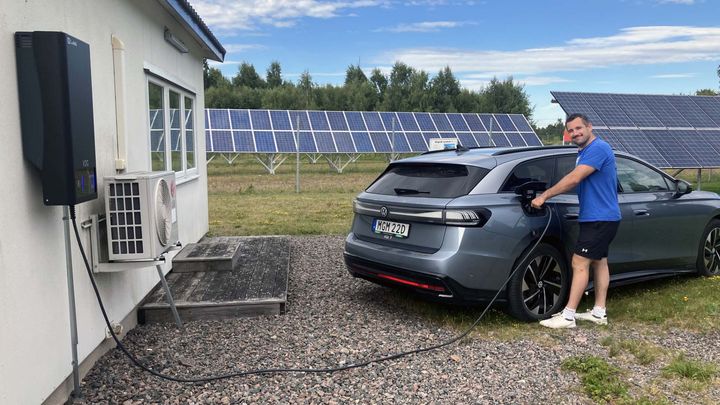 En man i blå t-shirt kopplar en laddkabel till en silverfärgad elbil utanför en vit byggnad, med solpaneler och grönska i bakgrunden.