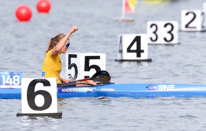 Kvinna i gul tröja paddlar förbi mållinjen i kajak med armen i luften, omgiven av nummerbojar.