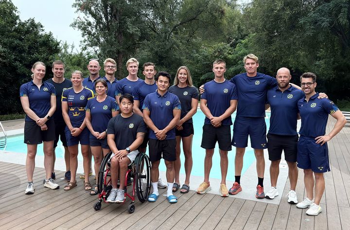 En grupp människor i sportkläder står samlade utomhus vid en pool. En person sitter i rullstol.