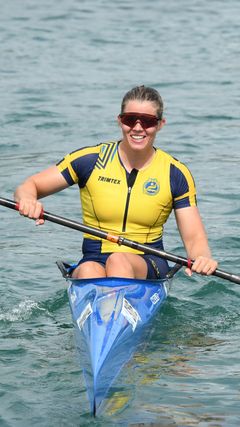 Kvinna i gult och blått träningsställ paddlar en blå kajak på vattnet.