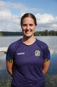 Person i mörkblå sporttröja står framför en sjö med skog och himmel i bakgrunden.