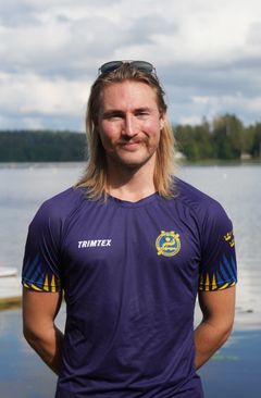 Person i mörkblå sporttröja står framför en sjö med skog och himmel i bakgrunden."