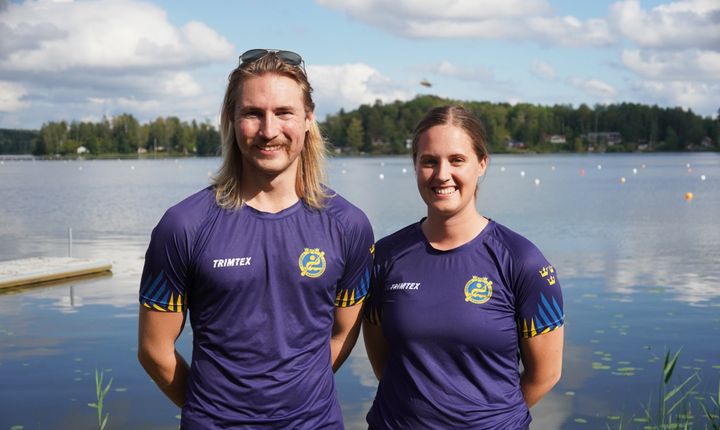 Två personer i blåa lagtröjor står vid en sjö med skog i bakgrunden.