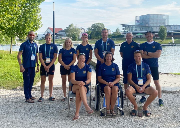 En gruppbild av den svenska truppen med tio personer klädda i blått, stående och sittande utomhus vid en sjö.