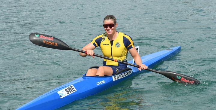 Bilden visar en kvinna i gul-blå tävlingsdräkt som paddlar en blå kajak i vattnet.