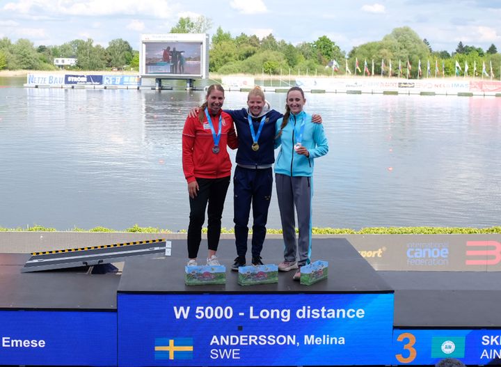 Tre kvinnliga idrottare står på prispallen vid en kanottävling. En av dem, Melina Andersson från Sverige, har vunnit guld.