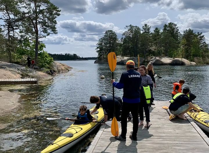 Folk paddlar kajak vid en träbrygga omgiven av träd och stenar.