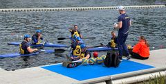En grupp män i kajaker får coachning av en person i sportkläder vid kanten av en vattenbana.