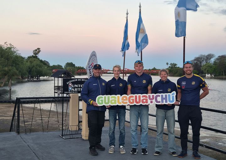 Fem personer i matchande kläder står vid en flodkant i skymning och håller upp en skylt med texten "Gualeguaychú", med argentinska flaggor och palmer i bakgrunden.