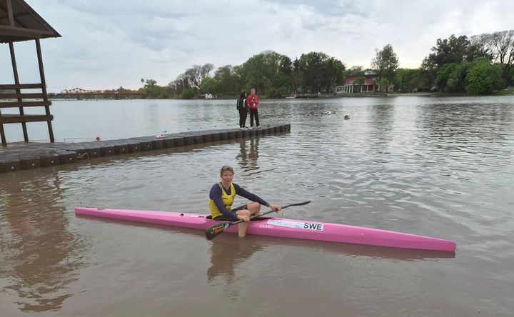 Kvinna i gul tröja paddlar en rosa kajak märkt "SWE" på en lugn flod, med en brygga och två personer i bakgrunden.