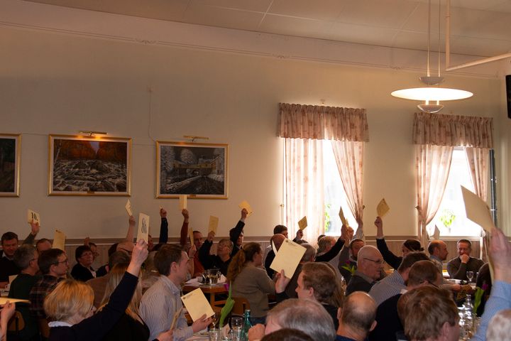 Människor vid stämman sitter i ett rum och håller upp papper, som om de röstar.