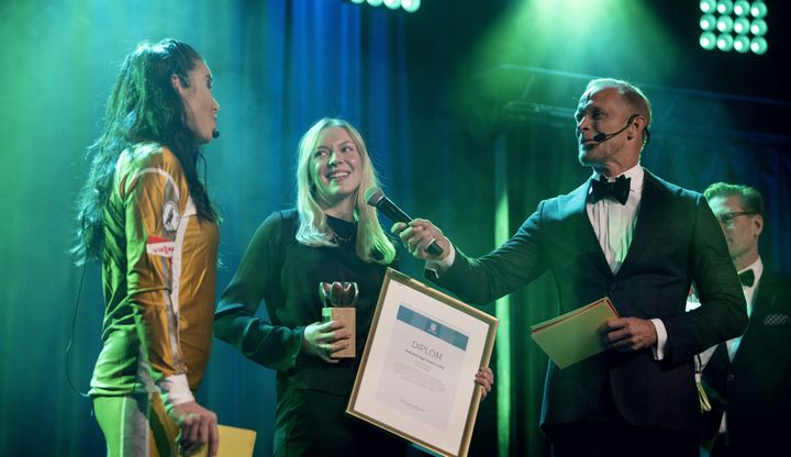 En person tar emot ett pris på scenen med två konferencierer.