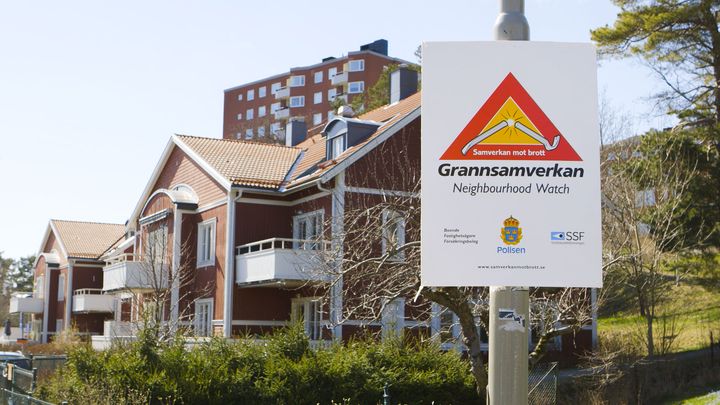 Skylt för Grannsamverkan framför röda hus och höghus i bakgrunden.