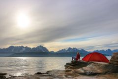 En person sitter vid ett stort vatten bredvid ett rött tält med berg i bakgrunden under en molnig himmel.