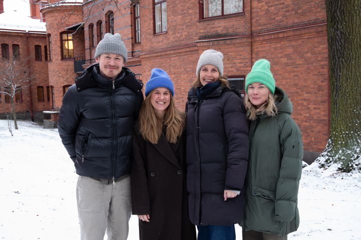Fyra personer står utomhus i vintermiljö framför en tegelbyggnad. De bär varma kläder och mössor.