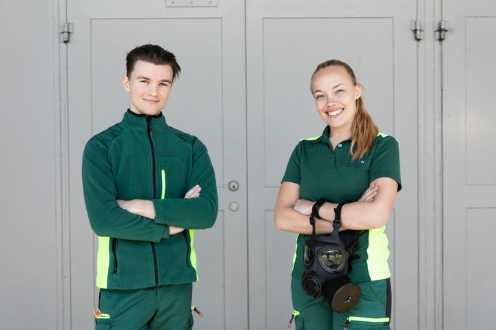 Tv%E5%20personer%20i%20gr%F6na%20arbetskl%E4der%20st%E5r%20leende%20med%20armarna%20i%20kors%20framf%F6r%20en%20ljusgr%E5%20v%E4gg.