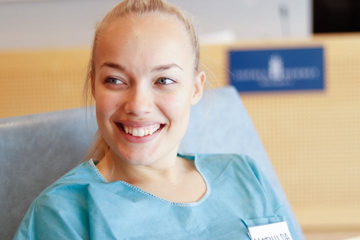 En%20leende%20person%20med%20blont%20h%E5r%20sitter%20i%20en%20stol%20kl%E4dd%20i%20en%20bl%E5%20topp.